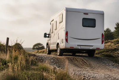 Vue arrière trois-quarts d’un camping-car montant une petite cassure de pente sur un chemin gravillonné, avec porte-à-faux visible