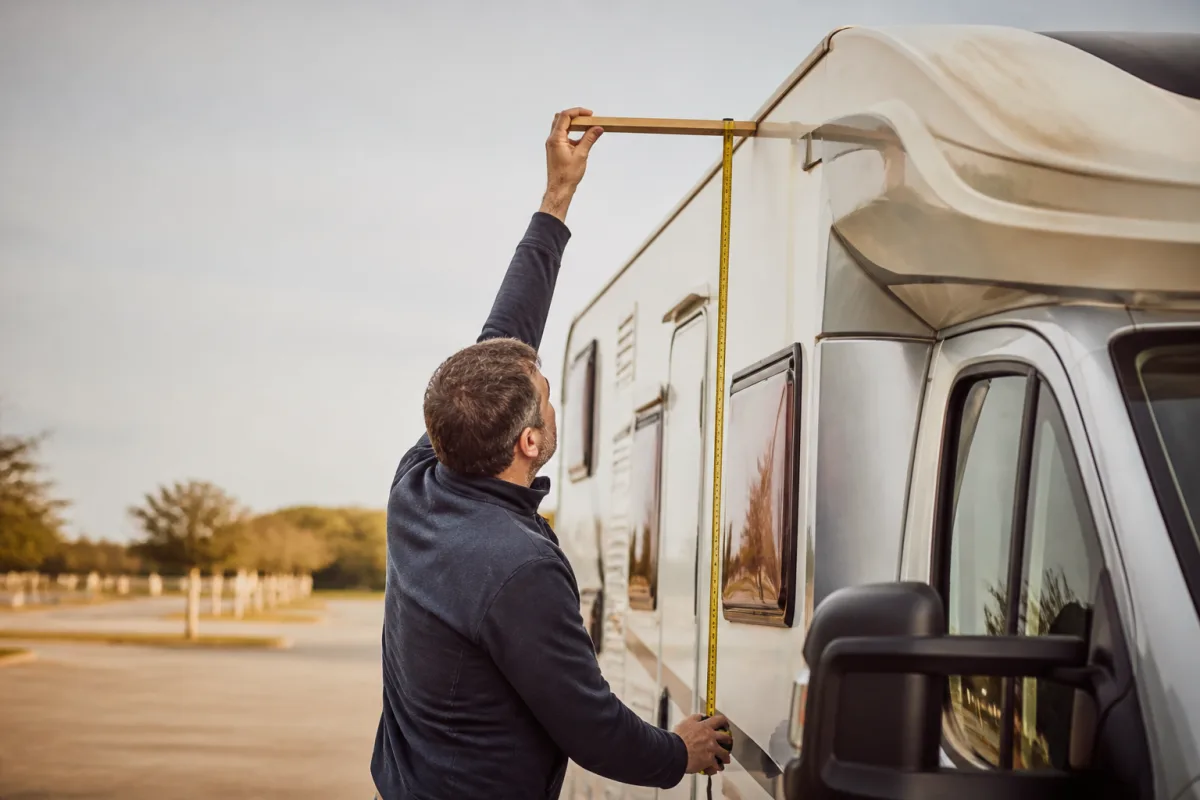 Main mesurant la hauteur d'un camping‑car avec un mètre ruban et une planchette, véhicule sur parking plat